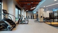 This contemporary fitness center is designed to deliver a complete workout experience within a spacious, single-floor layout. The facility is immediately distinguished by its bold architectural ceiling, featuring a rich terracotta-red paneled grid accented by sleek linear lighting and recessed spotlights that illuminate the space with a bright, energizing ambiance.

Large floor-to-ceiling windows line the perimeter walls, flooding the interior with natural light and offering open views of the surrounding urban streetscape. The cardio zone is particularly well-appointed, housing a full row of treadmills equipped with individual media screens, along with a selection of elliptical trainers and stationary bikes — all positioned to take advantage of the outdoor views.

Beyond cardio, the facility offers a robust strength training section, including cable and resistance machines, free weights with a dedicated dumbbell rack, and a variety of functional training equipment. Wall-mounted televisions are strategically placed throughout the space, ensuring entertainment is accessible from multiple workout zones.

The flooring transitions between durable light-toned carpet tile and wood-finish surfaces, defining separate zones while maintaining a cohesive aesthetic. The overall atmosphere is clean, modern, and professional, making this fitness center a well-rounded facility suited for a wide range of fitness routines.