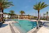 Located at 100 Inspire Way in St. Augustine, Florida, this outdoor amenity area serves as a vibrant centerpiece for the surrounding residential community. The space features two generously sized swimming pools with crystal-clear turquoise water, complemented by a dedicated spa, all finished with clean, light-toned paver decking that extends throughout the area.

Tall, mature palm trees are strategically planted throughout the space, providing both shade and a tropical ambiance that enhances the Florida lifestyle experience. The pool deck is well-appointed with a variety of outdoor lounge chairs, chaise lounges, and cushioned seating arrangements, offering residents ample space to relax and socialize. Market umbrellas are positioned throughout to provide additional shade.

The layout of the pool area is thoughtfully designed, with distinct zones for active swimming, lounging, and social gathering. A wrought-iron perimeter fence encloses the area, ensuring safety while maintaining open sightlines to the surrounding community and a scenic pond beyond. Views of the neighboring residential buildings and natural Florida landscape frame the space, contributing to a cohesive community atmosphere. Under the abundant Florida sunshine and clear blue skies, this outdoor retreat delivers a resort-style experience year-round.