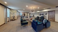 This well-appointed single-floor communal space is thoughtfully organized into several functional zones, each designed to encourage socializing and relaxation. The layout flows seamlessly from a generously sized lounge area to a more casual gathering section, offering flexibility for both intimate conversations and larger group activities.

The main lounge area is anchored by a pair of striking sputnik-style chandeliers that cast a warm, inviting glow over an arrangement of plush navy and teal velvet seating, including sofas and tufted accent chairs with gold-toned frames. A neutral, striped carpet grounds the space while complementing the rich jewel-toned furnishings. Framed artwork and decorative vases add curated touches throughout.

Adjacent to the lounge, a casual dining and activity area features round tables with modern swivel chairs in teal, set beneath recessed lighting and a complementary chandelier. Patterned drapery frames the windows, introducing soft natural light.

Near the entrance, a dedicated media corner with dark walls and theater-style seating provides an intimate screening experience. The overall ambiance is sophisticated yet comfortable, blending contemporary design elements with warm, welcoming details.