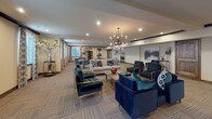 This well-appointed single-floor communal space is thoughtfully organized into several functional zones, each designed to encourage socializing and relaxation. The layout flows seamlessly from a generously sized lounge area to a more casual gathering section, offering flexibility for both intimate conversations and larger group activities.

The main lounge area is anchored by a pair of striking sputnik-style chandeliers that cast a warm, inviting glow over an arrangement of plush navy and teal velvet seating, including sofas and tufted accent chairs with gold-toned frames. A neutral, striped carpet grounds the space while complementing the rich jewel-toned furnishings. Framed artwork and decorative vases add curated touches throughout.

Adjacent to the lounge, a casual dining and activity area features round tables with modern swivel chairs in teal, set beneath recessed lighting and a complementary chandelier. Patterned drapery frames the windows, introducing soft natural light.

Near the entrance, a dedicated media corner with dark walls and theater-style seating provides an intimate screening experience. The overall ambiance is sophisticated yet comfortable, blending contemporary design elements with warm, welcoming details.