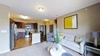 This single-floor residence offers a thoughtfully designed open-concept layout that integrates the kitchen, dining, and living areas into one cohesive and functional space. The kitchen is outfitted with dark espresso cabinetry, stainless steel appliances including a range and microwave, and a central island with bar seating and a stone-style countertop — ideal for casual dining or meal preparation. Rich hardwood-style flooring anchors the kitchen and dining zone, while plush carpet transitions seamlessly into the living area, adding warmth and comfort underfoot. The living space is generously proportioned, featuring neutral gray walls that provide a versatile backdrop for the contemporary furnishings. A large framed artwork in bold yellow and gray tones adds a striking accent to the room. The area is furnished with a comfortable sofa adorned with decorative pillows, an accent chair, and a round white coffee table, creating an inviting and cohesive arrangement. Flush-mount ceiling lighting provides consistent illumination throughout. Interior doors lead to additional private spaces, and the overall ambiance of the unit is modern, clean, and well-coordinated with a neutral palette accented by warm, earthy tones.