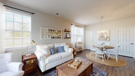 This single-floor residence offers a well-organized, open-concept layout with a warm coastal aesthetic throughout. The living area is generously proportioned, featuring rich hardwood-style flooring, neutral warm-toned walls, and multiple windows that fill the space with abundant natural light. A decorative geometric mirror, floating wood shelves, and sheer white curtains contribute to the room's airy character. A sliding vertical blind panel suggests access to an additional outdoor area.

The dining area flows naturally from the living space, anchored by a round wood table beneath an elegant gold candelabra chandelier. Natural light enters through large windows, and a decorative art piece adds personality to the neutral walls.

The kitchen is equipped with white cabinetry, granite countertops, and stainless steel appliances. A peninsula with bar seating provides additional prep space and casual dining functionality.

The bedroom is a calm retreat with soft carpeting, neutral walls, and a walk-in closet accessible through a dedicated doorway. Symmetrical nightstands and warm lighting enhance the room's comfortable atmosphere.

The bathroom features hardwood-style flooring, a single vanity with a clean white countertop, a mirrored medicine cabinet, and a full bathtub, maintaining the home's consistent, understated style.
