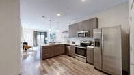 Located in the heart of Louisville, Kentucky, this modern 1-bedroom, 1-bathroom residence at 555 S Fourth St offers a thoughtfully designed open-concept layout. The combined kitchen and living area serves as the centerpiece of the home, featuring warm wood-look flooring and an abundance of natural light. The kitchen is outfitted with stainless steel appliances—including a French door refrigerator, electric range, built-in microwave, and dishwasher—complemented by wood-grain cabinetry, white quartz countertops, a subway tile backsplash, and a large island with seating for three. The living area is bright and welcoming, with access to a private balcony through a glass-panel door. The bedroom offers plush carpeting, a ceiling fan, and a window with city views, while an attached closet provides dedicated storage. The bathroom is finished with a wood-grain vanity, white quartz countertop, and a full tub-shower combination. Open shelving in the kitchen and a clean, neutral color palette throughout contribute to the contemporary aesthetic of this well-appointed residence in downtown Louisville.