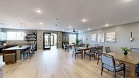 This well-appointed single-floor communal space offers a generously sized, open layout designed for group dining and social gatherings. The main dining area is anchored by multiple large wooden tables surrounded by upholstered chairs, all set atop light-toned hardwood flooring that runs throughout the space. Soft gray-green walls provide a calm, neutral backdrop, while a ceiling fan, recessed lighting, and wall sconces ensure the room is well-lit and comfortable. Curated artwork and lush indoor plants add warmth and character to the dining area.

A connected lounge nook features wicker chairs, a decorative area rug, and large windows with exterior views, allowing abundant natural light to filter in. French doors in this alcove open to a private balcony, further enhancing the airy ambiance. Decorative wall art accents the nook's intimate feel.

The fully equipped kitchen boasts rich dark wood cabinetry, granite countertops, stainless steel appliances, and a center island with bar seating. Open shelving provides display space, and a pendant chandelier adds a refined touch. Together, these spaces form a cohesive, functional, and elegantly designed environment ideal for communal living and entertaining.