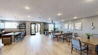 This well-appointed single-floor communal space offers a generously sized, open layout designed for group dining and social gatherings. The main dining area is anchored by multiple large wooden tables surrounded by upholstered chairs, all set atop light-toned hardwood flooring that runs throughout the space. Soft gray-green walls provide a calm, neutral backdrop, while a ceiling fan, recessed lighting, and wall sconces ensure the room is well-lit and comfortable. Curated artwork and lush indoor plants add warmth and character to the dining area.

A connected lounge nook features wicker chairs, a decorative area rug, and large windows with exterior views, allowing abundant natural light to filter in. French doors in this alcove open to a private balcony, further enhancing the airy ambiance. Decorative wall art accents the nook's intimate feel.

The fully equipped kitchen boasts rich dark wood cabinetry, granite countertops, stainless steel appliances, and a center island with bar seating. Open shelving provides display space, and a pendant chandelier adds a refined touch. Together, these spaces form a cohesive, functional, and elegantly designed environment ideal for communal living and entertaining.