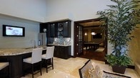 This single-floor space showcases a refined open-concept layout that seamlessly connects a wet bar area to a dedicated game room. The wet bar is outfitted with dark espresso cabinetry, a mosaic tile backsplash, and granite countertops, complemented by a stainless steel refrigerator and a breakfast bar with upholstered seating. The flooring throughout the main area features polished travertine tiles that lend a warm, luxurious feel. High ceilings amplify the sense of openness and grandeur. Adjacent to the bar, French-style glass-paneled doors open into a game room centered around a billiard table, finished with rich hardwood flooring and warm ambient lighting from stylish pendant fixtures. A wall-mounted flat-screen television and decorative wall sconces enhance the entertainment atmosphere of this secondary space. A large indoor plant adds a natural, organic element to the transition between areas. The overall ambiance is sophisticated and masculine, with a cohesive dark wood and neutral stone palette that creates an upscale, resort-inspired feel throughout the space.