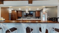 Located at 7200 Chadwood Lane in Dublin, Ohio, this single-story space offers a compelling blend of rustic architectural character and modern finishes. The main living area is defined by dramatic vaulted ceilings clad in rich, dark-stained wood planks, supported by substantial exposed beams that lend a warm, lodge-like atmosphere. A skylight overhead draws natural light into the space, complementing the large windows that frame outdoor views of the surrounding landscape. A floor-to-ceiling gray brick fireplace serves as the room's focal point, paired with a wall-mounted television above. Large-format tile flooring runs throughout, providing a clean, cohesive foundation.

A fully equipped galley-style kitchen features floor-to-ceiling rich walnut cabinetry, marble-style countertops, stainless steel appliances, and tile flooring consistent with the rest of the space. An adjacent bar area includes a thick wood countertop, built-in sink, additional cabinetry, and decorative pendant lighting — ideal for entertaining.

A glass-enclosed room provides a versatile, semi-private area visible from multiple vantage points throughout the layout. The overall ambiance balances warmth and sophistication, making this a well-appointed and functional space throughout.