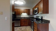 This single-floor space is thoughtfully designed to maximize functionality and comfort across its key areas. The kitchen serves as the centerpiece of the unit, showcasing a U-shaped layout with rich, medium-toned wood cabinetry extending from floor to ceiling, providing generous storage throughout. Dark granite countertops with subtle veining offer a striking contrast against the cabinetry and are paired with a full suite of stainless steel appliances, including a refrigerator, dishwasher, electric range, and over-the-range microwave. Under-cabinet lighting illuminates the workspace, while stone-look tile flooring adds durability and visual warmth. A flush-mount ceiling fixture provides ample overhead lighting. The kitchen is accessible from an adjacent hallway, which transitions seamlessly between rooms. The bathroom features a full tub-shower combination with cream-toned tile surround, a vanity with dark wood cabinetry, a white countertop, and matching stone-look tile flooring, creating a cohesive aesthetic. The bedroom is a calm, neutral space with soft carpeting, light gray walls, white panel doors, and clean white baseboards, offering a comfortable and versatile environment.