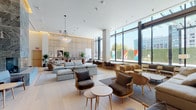 This expansive single-floor lounge showcases a refined contemporary design that balances openness with intimate seating areas. Soaring ceilings amplify the sense of volume throughout the space, while an extensive wall of floor-to-ceiling windows floods the interior with natural light and frames views of the surrounding urban landscape, including a vibrant colorful mural visible from the exterior terrace.

A commanding floor-to-ceiling marble fireplace serves as a dramatic architectural focal point, clad in richly veined gray stone and flanked by built-in shelving displaying curated books and décor. Opposite the fireplace, a slatted wood partition wall adds warmth and visual texture, subtly dividing the space into distinct zones without sacrificing the open, airy feel.

Light-toned hardwood flooring runs throughout, complementing a neutral palette of soft grays, warm beiges, and natural wood tones. Multiple seating arrangements feature modular sofas, rattan chairs, and woven accent chairs grouped around low tables, accommodating both social gatherings and quiet individual use. Tall potted tropical plants are strategically placed throughout, enhancing the organic, biophilic ambiance. A dedicated workstation area with bench seating along the window wall provides a functional workspace within this elegantly designed communal setting.