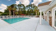 Located along W Highway 98 in Pensacola, Florida, this outdoor pool facility offers a well-appointed recreational retreat designed for relaxation and leisure. The centerpiece is a generously sized swimming pool with crystal-clear turquoise water, accessible via stainless steel handrails and a gradual entry staircase on one end, making it convenient for swimmers of all abilities. The expansive concrete pool deck surrounds the pool on all sides, providing ample space for lounging and socializing. Numerous chaise lounge chairs are arranged along the deck, offering prime spots to soak up the Florida sun. Shade options include vibrant red hammock-style shade sails strung between posts and blue market umbrellas positioned over seating areas, ensuring comfort throughout the day. A covered portico with classic white columns anchors one corner of the deck, adding a touch of architectural elegance to the outdoor setting. The entire area is enclosed by a traditional white picket fence, lending a clean, classic aesthetic. Tall pine trees, mature palms, and lush greenery form a natural backdrop, creating a sense of seclusion and privacy. A single-story clubhouse or pool house with a gabled roof, brick accents, and multiple French doors lines one side of the pool, providing easy indoor access to facilities.