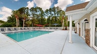 Located along W Highway 98 in Pensacola, Florida, this outdoor pool facility offers a well-appointed recreational retreat designed for relaxation and leisure. The centerpiece is a generously sized swimming pool with crystal-clear turquoise water, accessible via stainless steel handrails and a gradual entry staircase on one end, making it convenient for swimmers of all abilities. The expansive concrete pool deck surrounds the pool on all sides, providing ample space for lounging and socializing. Numerous chaise lounge chairs are arranged along the deck, offering prime spots to soak up the Florida sun. Shade options include vibrant red hammock-style shade sails strung between posts and blue market umbrellas positioned over seating areas, ensuring comfort throughout the day. A covered portico with classic white columns anchors one corner of the deck, adding a touch of architectural elegance to the outdoor setting. The entire area is enclosed by a traditional white picket fence, lending a clean, classic aesthetic. Tall pine trees, mature palms, and lush greenery form a natural backdrop, creating a sense of seclusion and privacy. A single-story clubhouse or pool house with a gabled roof, brick accents, and multiple French doors lines one side of the pool, providing easy indoor access to facilities.