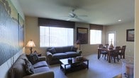 This single-floor residence features an open-concept living and dining area that maximizes the sense of space and functionality. The living area is furnished with a plush sectional sofa and matching loveseat arranged around a dark wood coffee table, creating a comfortable and inviting seating arrangement. A ceiling fan is centrally mounted overhead, providing both practical air circulation and a subtle decorative element.

Natural light fills the room through multiple windows dressed with vertical blinds and dark valances, offering both style and light control. The neutral-toned walls and carpeted flooring contribute to a warm, cohesive ambiance throughout the space. A framed landscape painting serves as a focal point on the main wall, adding a touch of color and character to the room.

The dining area is seamlessly integrated into the same open space, featuring a dark wood table paired with matching chairs, making it ideal for casual meals or entertaining. Soft accent lighting from a floor lamp complements the natural light, enhancing the overall warmth of the interior. The layout is practical and well-proportioned, making efficient use of the available space.