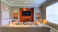 This single-story lounge space combines contemporary design with warm, inviting tones. The focal point of the room is a recessed orange accent wall housing a large wall-mounted flat-screen television, flanked by two matching landscape paintings that echo the warm earth-toned color palette. Recessed lighting illuminates the space from above, complementing the crown molding that frames the ceiling and adds a refined architectural detail.

The seating arrangement is thoughtfully organized around a central rectangular coffee table, which serves as a functional gathering point. A generous neutral-toned sofa anchors the foreground, while two rust-colored armchairs are positioned near the accent wall, creating an intimate conversation area. A mustard-yellow lounge chair adds a complementary pop of color to the right side of the room. A cushioned bench provides additional flexible seating near the perimeter.

The flooring features a subtly patterned carpet in neutral tones that ties the warm and cool color elements together cohesively. Overall, the space exudes a polished, community-oriented ambiance suited for relaxation or casual gatherings.