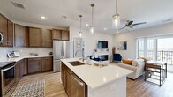 This single-floor residence on Legends Drive in Champaign, Illinois, is designed with a modern, open-concept layout that seamlessly connects the main living areas. The kitchen serves as a central hub, featuring rich medium-toned cabinetry, a full suite of stainless steel appliances, subway tile backsplash, and a generously sized island with quartz countertops, an undermount sink, and seating for casual dining. Pendant lighting illuminates the island, adding a refined touch to the space.

The kitchen flows directly into the living room, which is bright and airy, anchored by a sleek linear fireplace beneath a wall-mounted television. A sliding glass door opens onto a private balcony, offering open views of the surrounding landscape, including a pool area visible in the distance. Light wood-style flooring runs throughout the main living areas, while plush carpet adds warmth to the bedrooms.

The bedrooms are comfortably sized with ample natural light provided by large windows. Ceiling fans contribute to comfort throughout. The bathrooms are finished with wood-toned vanities, light countertops, and tub-shower combinations, maintaining a consistent, cohesive design palette across the entire residence.