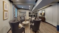 This thoughtfully designed communal lounge space exudes a refined, upscale ambiance throughout. The interior is anchored by warm hardwood-style flooring that runs continuously across the open layout, complemented by bold dark wood beam accents that frame the ceilings and add architectural character. Recessed lighting illuminates the space evenly, enhancing the inviting atmosphere.

The main seating area showcases a curated arrangement of charcoal grey leather wingback chairs with brass nailhead trim, gathered around a marble-topped cocktail table with a gold base — a striking focal point that balances elegance with comfort. Nearby, a rich dark wood credenza topped with gold table lamps and decorative florals adds warmth and depth to the space.

Large windows dressed with patterned drapery in geometric motifs allow natural light to filter in while providing a stylish visual frame to the outdoors. Arched architectural details and structural columns contribute a classic, refined touch to the overall design.

A wrought iron railing lines a staircase area, connecting the lounge to adjacent corridors and spaces, reinforcing the sense of an open, interconnected environment. A secondary seating vignette features plush navy blue accent chairs, a gilded sunburst mirror, and framed wall art, completing the cohesive and polished aesthetic of this welcoming shared space.
