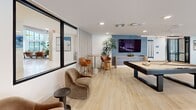 Located at 20700 North 55th Street in Phoenix, AZ, this single-floor amenity lounge offers a thoughtfully designed open-concept space that seamlessly blends a fully equipped kitchen with an inviting living and recreation area. The kitchen is anchored by a striking elongated island with a sculpted wood-paneled base and white quartz countertop, surrounded by warm leather barstools. Globe pendant lights hang overhead, complementing the wood-toned cabinetry, white countertops, stainless steel appliances, and an eye-catching textured accent wall with a wall-mounted TV. Light hardwood flooring runs throughout, lending warmth and cohesion to the entire space. The living area transitions effortlessly into a recreation zone, where a full-size billiards table takes center stage beneath a brass pendant light fixture. A blue textured feature wall with a mounted TV anchors this side of the room, flanked by a tall indoor palm that adds a natural, relaxed touch. Plush curved lounge chairs and bistro-height bar seating create intimate gathering spots throughout. Floor-to-ceiling black-framed glass partitions and windows flood the space with natural light while maintaining a sleek, modern aesthetic. The overall ambiance is polished yet approachable, ideal for community gathering and leisure.
