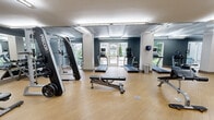 This fitness center offers a spacious, single-floor layout designed to accommodate a full range of workout activities. The open floor plan is finished with light wood-tone flooring throughout, creating a clean and energizing environment. Structural columns are positioned throughout the space, subtly dividing functional zones without disrupting the overall flow.

The cardio section is well-appointed with multiple treadmills, elliptical trainers, and a stair climber, each equipped with individual monitors. Large windows and glass doors along the perimeter flood the space with natural light and offer views of the surrounding greenery, enhancing the workout atmosphere.

The strength training area is comprehensively stocked, featuring a multi-station cable and weight machine, a dedicated dumbbell rack with a full range of free weights, adjustable weight benches, and a plate-loaded barbell station. Additional equipment such as a cable crossover system and adjustable benches provide versatility for varied training routines.

Wall-mounted mirrors along key sections of the space visually expand the room and allow users to monitor their form. Overhead fluorescent lighting ensures the entire floor is evenly and brightly illuminated. A shelving unit along one wall provides convenient storage. The overall ambiance is modern, functional, and well-maintained, making it a thorough facility for both cardio and strength training needs.