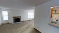 This single-floor space offers a clean, functional layout with classic residential finishes throughout. The main living area is a standout feature, centered around a tile-surround fireplace with a wood mantel that adds warmth and character to the room. Plush wall-to-wall carpeting runs throughout the space, contributing to a comfortable and inviting atmosphere. A large window allows generous natural light to fill the room, creating a bright and open feel. An elegant chandelier hangs nearby, providing both ambient and decorative lighting that enhances the overall aesthetic. The walls are finished in a crisp white, complementing the neutral tones of the carpet and contributing to an airy, spacious ambiance. A textured ceiling adds a subtle traditional detail. Baseboard heating lines the perimeter, offering efficient and consistent warmth throughout the colder months. The open arrangement of the living space allows for flexible furniture placement, making it well-suited for a variety of uses. Overall, this space combines practical features with comfortable, understated design elements that create a welcoming and livable environment.