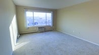 This single-floor space features a generously sized main living area finished with wall-to-wall carpeting in a neutral tone, providing a comfortable and quiet underfoot feel. The room is anchored by a large picture window dressed with simple white curtains, allowing natural light to fill the interior and offering views of the surrounding exterior. Beneath the window, a wall-mounted air conditioning unit sits alongside a baseboard heating system, ensuring year-round climate control throughout the space. The walls are painted in a soft, warm beige tone, contributing to a clean and understated aesthetic that serves as a versatile backdrop for a variety of furnishing styles. The open layout of the living area allows for flexible furniture arrangement, making it easy to configure the space according to personal needs. White baseboards provide a subtle contrast against the carpeted floor, adding a modest finishing touch to the room's overall look. The space presents a straightforward and functional design, well-suited for comfortable everyday living.