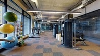 This fitness center offers a spacious, single-floor layout designed to accommodate a comprehensive workout experience. The space is defined by its industrial aesthetic, featuring an exposed concrete ceiling with visible ductwork and ventilation systems, illuminated by rows of sleek linear pendant lighting that runs the length of the room. A wall of floor-to-ceiling windows lines one side of the facility, flooding the interior with abundant natural light and creating an open, energizing atmosphere. The flooring consists of a durable, multi-toned carpet tile in a checkered pattern of warm and cool tones, providing both comfort and visual interest underfoot. Along the windowed wall, a lineup of cardio equipment — including treadmills, ellipticals, and rowing machines — is arranged to take full advantage of the natural light and views beyond. On the opposite side, a series of large, professional-grade strength training machines and cable systems are positioned to create a dedicated resistance training zone. The combination of ample space between equipment, high ceilings, and generous lighting contributes to an open and functional environment well-suited for a broad range of fitness activities.