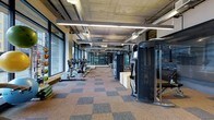 This fitness center offers a spacious, single-floor layout designed to accommodate a comprehensive workout experience. The space is defined by its industrial aesthetic, featuring an exposed concrete ceiling with visible ductwork and ventilation systems, illuminated by rows of sleek linear pendant lighting that runs the length of the room. A wall of floor-to-ceiling windows lines one side of the facility, flooding the interior with abundant natural light and creating an open, energizing atmosphere. The flooring consists of a durable, multi-toned carpet tile in a checkered pattern of warm and cool tones, providing both comfort and visual interest underfoot. Along the windowed wall, a lineup of cardio equipment — including treadmills, ellipticals, and rowing machines — is arranged to take full advantage of the natural light and views beyond. On the opposite side, a series of large, professional-grade strength training machines and cable systems are positioned to create a dedicated resistance training zone. The combination of ample space between equipment, high ceilings, and generous lighting contributes to an open and functional environment well-suited for a broad range of fitness activities.