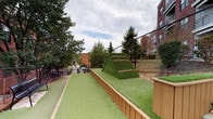 Del Ray Central's amenity space combines a well-designed outdoor turf area with functional interior circulation elements. The outdoor area is surfaced entirely in artificial turf and features a distinctive pyramid-shaped climbing structure covered in synthetic grass with integrated wooden step ledges, serving as a focal point of the space. Raised planter beds with wood composite borders line the perimeter, adding a structured, landscaped character. The area is enclosed by chain-link fencing, providing security while maintaining open sightlines to surrounding mature trees and neighboring brick buildings. The layout offers a linear pathway alongside the pyramid feature, creating a defined activity corridor.

Interior access is provided through a utilitarian stairwell constructed with painted concrete block walls and concrete flooring. Metal handrails guide occupants along the stairs, which connect levels and lead to exit doors equipped with push-bar hardware. Signage within the stairwell directs users to rooftop access and level exits, reflecting a multi-level building configuration. The stairwell's design is straightforward and functional, emphasizing safety and circulation efficiency. Together, the outdoor and interior elements form a cohesive amenity package characteristic of a modern urban mixed-use development.