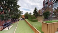 Del Ray Central's amenity space combines a well-designed outdoor turf area with functional interior circulation elements. The outdoor area is surfaced entirely in artificial turf and features a distinctive pyramid-shaped climbing structure covered in synthetic grass with integrated wooden step ledges, serving as a focal point of the space. Raised planter beds with wood composite borders line the perimeter, adding a structured, landscaped character. The area is enclosed by chain-link fencing, providing security while maintaining open sightlines to surrounding mature trees and neighboring brick buildings. The layout offers a linear pathway alongside the pyramid feature, creating a defined activity corridor.

Interior access is provided through a utilitarian stairwell constructed with painted concrete block walls and concrete flooring. Metal handrails guide occupants along the stairs, which connect levels and lead to exit doors equipped with push-bar hardware. Signage within the stairwell directs users to rooftop access and level exits, reflecting a multi-level building configuration. The stairwell's design is straightforward and functional, emphasizing safety and circulation efficiency. Together, the outdoor and interior elements form a cohesive amenity package characteristic of a modern urban mixed-use development.