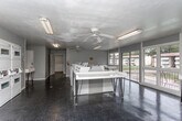 Located at 464 N Oakley Dr in Columbus, Georgia, this single-floor community laundry facility is designed to serve residents with a practical and functional layout. The space features a generous row of top-loading washing machines lining one side of the room, complemented by stacked dryer units positioned along the adjacent wall, providing ample laundry capacity for a large number of users. Folding tables are conveniently placed throughout the room, offering dedicated surfaces for sorting and folding laundry. The interior is finished with yellow-green painted walls and dark flooring, giving the space a clean, utilitarian appearance. Overhead fluorescent lighting ensures the room is well-lit throughout. Large windows and a sliding glass door at one end of the facility flood the space with natural light and provide views of the surrounding grounds and neighboring buildings. A fire extinguisher is mounted on the wall, and safety signage is posted throughout, reflecting attention to occupant safety. An additional room accessible through an interior doorway appears to house more laundry equipment. The facility's open, spacious configuration allows for comfortable movement between machines and folding areas, making it a well-equipped and accessible amenity for the community.