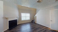 This two-story residence is finished with dark hardwood-style flooring that runs consistently throughout both levels, contributing to a cohesive and polished aesthetic. The main living area on the lower floor features vaulted-style ceilings, a fireplace with a mantel, and a window fitted with blinds that allows natural light to fill the space. A staircase along one wall leads to the upper level, creating an open vertical feel in the living room.

The galley-style kitchen is well-appointed with dark espresso cabinetry on both sides, a mosaic tile backsplash, stainless steel appliances including a range and microwave, and light-toned countertops. A double basin sink sits beneath upper cabinets on one side, and the layout maximizes storage and counter space efficiently. The kitchen connects to the dining area, which features a ceiling fan and access to additional rooms.

The upper level, reached via the interior staircase, includes a generously sized bedroom with a ceiling fan, windows on two sides, and a sliding door with vertical blinds. An en-suite bathroom features dark vanity cabinetry, a framed mirror, a well-lit vanity with light countertops, and an adjacent shower/tub combination. A textured popcorn ceiling is present throughout the home.