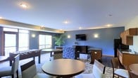 This single-floor community lounge is designed to offer a relaxed and social environment for its occupants. The space is defined by its cool blue walls, which lend a calm and inviting atmosphere throughout. Recessed ceiling lighting is complemented by warm wall sconces positioned around the room, creating a layered and cozy ambiance even as natural light filters in through a large window overlooking a greenery-filled exterior view.

The layout accommodates a variety of activities and social settings. Round dining-height tables with upholstered chairs are arranged throughout, offering comfortable spots for gathering or casual meals. A foosball table adds a recreational element, making the space well-suited for leisure and entertainment. Toward one end of the room, a wall-mounted flat-screen television is flanked by a media console and a bar-height counter with stools, creating a dedicated area for watching and unwinding.

Seating variety continues with a pair of patterned blue accent chairs, adding a pop of decorative flair that ties into the room's overall color palette. A tall potted tropical plant near the window introduces a natural, refreshing element to the interior. The patterned carpeting grounds the space and contributes to its warm, community-focused character. Overall, this lounge is thoughtfully appointed to serve as a versatile and comfortable gathering space.
