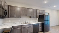 Located at 1202 N Main St in Nixa, Missouri, this single-floor unit offers a clean, modern aesthetic throughout its thoughtfully designed layout. The kitchen serves as a functional centerpiece, showcasing rich dark-stained cabinetry with sleek brushed-nickel hardware, marble-look countertops, and a full suite of black appliances including a range, over-the-range microwave, dishwasher, and refrigerator. Light wood-look flooring runs through the kitchen and living areas, lending warmth and continuity to the space. The living area features a sliding glass door fitted with vertical blinds, allowing for natural light and outdoor access. Bedrooms are carpeted for comfort and are served by generous closet space, including louvered bifold doors and a rod-and-shelf closet system. A ceiling fan adds functionality to the bedroom area. The bathrooms are well-appointed with dark wood vanities, marble-look countertops, brushed-nickel fixtures, and bright vanity lighting above large mirrors. The overall color palette—neutral walls paired with warm wood tones and stone-look surfaces—creates a cohesive, contemporary feel throughout the unit.
