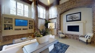 This remarkable single-floor space makes an immediate impression with its dramatic architectural character. Soaring ceilings anchored by rich, dark wood ceiling beams create a grand sense of vertical space, while exposed brick columns with arched detailing add warmth and historic charm throughout. Banks of large clerestory windows line the upper walls, drawing in generous amounts of natural light and offering views of the surrounding mature trees.

The main living area is elegantly appointed, featuring a sleek, light-toned stone countertop surface at the foreground that serves as a central focal point. A built-in shelving unit houses a wall-mounted flat-screen television alongside curated décor, flanked by a fireplace with a classic white surround and brick detailing. The seating arrangement includes stylish accent chairs, a patterned area rug with a bold dotted motif, and carefully selected greenery and botanical arrangements that bring life and texture to the space.

Warm-toned walls complement the brick and wood elements, while light hardwood flooring ties the entire space together cohesively. A statement chandelier overhead adds a touch of elegance. The overall ambiance is one of sophisticated comfort — blending rustic architectural elements with refined, contemporary furnishings for a welcoming and visually striking environment.