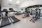 This fitness center in Huntsville, Alabama, is designed to support a comprehensive workout experience within a single-floor layout. The space features vaulted ceilings that lend an open, airy feel, complemented by light wood-style flooring throughout the main exercise areas. Ceiling fans provide consistent air circulation, while a combination of overhead lighting and wall-mounted fixtures ensures the space remains well-lit during use.

The cardio section is equipped with multiple treadmills, elliptical machines, and a stationary bike, arranged to allow comfortable spacing between users. A wall-mounted flat-screen television is positioned for easy viewing during cardio sessions. Adjacent to the cardio area, a dedicated strength training zone features a full dumbbell rack with varying weights, a kettlebell, and an adjustable workout bench, all set on protective rubber flooring mats.

A large wall-to-wall mirror spans one side of the strength training area, visually expanding the space and allowing users to monitor their form. The design incorporates a bold black accent wall, adding a modern, motivational aesthetic to the overall environment. Natural light filters in through multiple windows fitted with horizontal blinds, further enhancing the bright and energetic atmosphere of the facility.