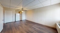 This one-bedroom, one-bathroom unit at 5300 Hawthorne Rd in Roanoke, Virginia offers a functional and efficiently arranged layout across a single floor. The living area is open and straightforward, featuring rich dark hardwood-style flooring that runs throughout the main spaces, creating a cohesive and clean aesthetic. Drop tile ceilings with ceiling fans are present in the main rooms, providing both air circulation and overhead lighting. The paneled walls in soft neutral tones lend a understated, uniform character to the space. Natural light filters in through windows fitted with blinds, offering views of the surrounding area. The kitchen is compact and practical, equipped with a full-size refrigerator and wooden cabinetry. The spacious bedroom includes its own ceiling fan, a window for natural light, and baseboard heating, ensuring year-round comfort. A dedicated closet provides useful storage. The bathroom is designed with accessibility in mind, featuring grab bars and a step-in shower with a built-in seat and non-slip flooring. The closet area features vinyl tile flooring and painted concrete block walls, adding a sturdy, no-frills utility space. Overall, the unit presents a clean, practical living environment well-suited for those seeking straightforward accommodations in Roanoke.