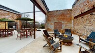 This distinctive single-level space in South Carolina is defined by its striking industrial character, anchored by soaring exposed brick walls that bear the patina of age, with layers of weathered paint adding depth and texture throughout. The open-air courtyard design creates a dynamic environment that blends historical architecture with modern amenities.

The space is organized into several functional zones. A lounge area features teak-framed sofas and chairs with cream cushions and green accent pillows, arranged around large cantilever umbrellas for shade. Adjacent to this is a fire pit gathering area, where a substantial concrete bowl fire pit is encircled by rustic wooden slatted chairs, perfect for intimate conversation. A bar-height dining area with wicker seating and a market umbrella provides an additional social hub near the perimeter fence, which opens to surrounding greenery and views beyond.

A dedicated recreation zone houses a full-size Kettler ping-pong table set against the weathered brick backdrop. Structural steel beams and exposed conduit piping add to the industrial aesthetic, while French doors provide interior access points. The polished concrete flooring unifies all zones, and the open sky above floods the entire space with natural light, creating a seamless indoor-outdoor atmosphere.