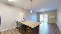 Located at 1602 Belsly Blvd in Moorhead, MN, this one-bedroom, one-bathroom unit offers a well-designed, single-floor layout that maximizes functionality and comfort. The open-concept kitchen and living area serves as the heart of the home, anchored by a peninsula-style kitchen island topped with stone-look countertops and fitted with a double stainless steel sink and sleek pendant lighting. Dark espresso cabinetry with brushed chrome hardware provides ample storage, complemented by black appliances including a refrigerator, range, microwave, and dishwasher. Wood-look vinyl plank flooring runs throughout the main living area, adding warmth and durability. A sliding glass door with vertical blinds floods the living room with natural light and opens to an outdoor patio area. The bedroom features plush carpet flooring, a window with blinds offering outdoor views, and baseboard heating. The full bathroom is well-appointed with a stone-look vanity countertop matching the kitchen, dark wood cabinetry, a tub/shower combo, and tile flooring. A dedicated in-unit laundry room with tile flooring and shelving rounds out this thoughtfully designed home.
