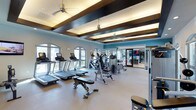 This fitness center offers a thoughtfully designed workout environment that balances functionality with an upscale aesthetic. The space is defined by its striking coffered ceiling, accented with dark wood beams and recessed lighting, which adds architectural character while ceiling fans maintain comfortable airflow throughout. Large arched windows line one wall, flooding the interior with natural light and providing views of the surrounding greenery, creating an inviting atmosphere for exercise. The main floor is dedicated to a generous selection of cardio equipment, including a row of treadmills and elliptical machines, all outfitted with individual screens for entertainment during workouts. Strength and functional training equipment occupies the central area, offering versatility for a range of exercise routines. The accent wall, painted in a vibrant teal, adds a bold visual element that energizes the space. A mirrored studio area, accessible through glass doors, provides a dedicated zone for stretching, group classes, or independent training. The durable, easy-to-maintain flooring throughout supports heavy equipment use while contributing to the clean, polished look of the overall space.