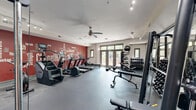 The fitness center at Chapel Hill North, located at 200 Perkins Dr in Chapel Hill, NC, is a thoughtfully designed exercise space offering a comprehensive range of workout options. The room features durable rubber flooring throughout, providing a safe and functional surface for all types of training.

The cardio zone is anchored by a bold red accent wall emblazoned with a motivational fitness-themed word mural prominently featuring "Chapel Hill North." This area houses treadmills, an elliptical, and a stair climber, all positioned beneath a ceiling fan and track lighting for comfortable workouts. 

The free weight section includes a full dumbbell rack and adjustable benches, positioned near large glass double doors that open to an outdoor patio, allowing natural light and views of surrounding greenery to fill the space. A floor-to-ceiling mirror along one wall enhances the sense of openness and aids in monitoring form.

The strength training area features a multi-functional cable machine and additional weight equipment, complemented by a glass partition wall that adds visual depth to the room. Track lighting throughout ensures consistent, well-distributed illumination across all workout zones. The space is efficiently laid out to accommodate a variety of fitness routines, making it a versatile and practical facility for residents.