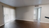 This single-story, one-bedroom, one-bathroom unit offers a functional and well-organized layout designed to maximize every square foot of available space. The living room is a bright, open area featuring dark hardwood-style flooring, neutral gray walls, and a large sliding glass door fitted with vertical blinds that allows natural light to fill the room. The galley-style kitchen is efficiently arranged with white cabinetry, dark granite-look countertops, and black appliances including a refrigerator and electric range, complemented by a stainless steel sink and in-unit washer and dryer connections visible toward the rear. The bedroom is carpeted and comfortably sized, equipped with a ceiling fan and a window with blinds that brings in natural light. A dedicated walk-in closet with wire shelving provides generous storage directly off the bedroom. The bathroom features a full-size tub and shower combo with clean white surround panels, a toilet, and dark wood-style flooring that ties back to the main living areas. Neutral tones throughout the unit, combined with consistent flooring choices and clean white baseboards, create a cohesive and practical living environment.