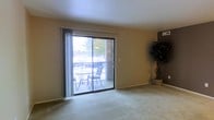 This single-floor residence offers a well-organized layout designed for comfortable everyday living. The living area is carpeted and bathed in natural light, courtesy of a large sliding glass door that opens onto a private balcony. The balcony, framed by a brick exterior wall and metal railing, provides an outdoor seating area with views of the surrounding landscape, making it a pleasant extension of the interior living space. Vertical blinds on the sliding door allow for adjustable light control and privacy. The neutral-toned walls and light carpeting throughout the living area contribute to a clean, understated aesthetic.

The kitchen is efficiently designed with a galley-style layout enclosed by an arched overhead structure. White cabinetry lines the upper walls, providing ample storage, while white appliances—including a gas range, refrigerator, and countertop microwave—equip the space for daily use. Track lighting overhead ensures the kitchen is well-lit. A wraparound breakfast bar separates the kitchen from the adjacent area and offers casual seating with two white bar stools, creating a convenient dining or workspace. The overall ambiance of the residence is functional, tidy, and inviting.