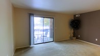 This single-floor residence offers a well-organized layout designed for comfortable everyday living. The living area is carpeted and bathed in natural light, courtesy of a large sliding glass door that opens onto a private balcony. The balcony, framed by a brick exterior wall and metal railing, provides an outdoor seating area with views of the surrounding landscape, making it a pleasant extension of the interior living space. Vertical blinds on the sliding door allow for adjustable light control and privacy. The neutral-toned walls and light carpeting throughout the living area contribute to a clean, understated aesthetic.

The kitchen is efficiently designed with a galley-style layout enclosed by an arched overhead structure. White cabinetry lines the upper walls, providing ample storage, while white appliances—including a gas range, refrigerator, and countertop microwave—equip the space for daily use. Track lighting overhead ensures the kitchen is well-lit. A wraparound breakfast bar separates the kitchen from the adjacent area and offers casual seating with two white bar stools, creating a convenient dining or workspace. The overall ambiance of the residence is functional, tidy, and inviting.