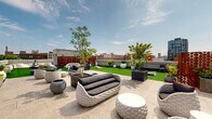 Located in Philadelphia's historic Old City neighborhood, this generously sized rooftop terrace offers an exceptional outdoor experience above the city. The space is thoughtfully divided into distinct zones, combining large-format stone tile patio areas with lush artificial turf lawns that create a vibrant, park-like atmosphere high above street level. Throughout the terrace, an extensive collection of planters — ranging from sculptural round vessels to tall rectangular containers — showcases a variety of trees, shrubs, and flowering plants, bringing rich greenery to the urban setting. Multiple seating arrangements are distributed across the space, featuring contemporary outdoor sofas, lounge chairs, accent chairs, and sun loungers, providing versatile areas for relaxation and social gatherings. A built-in outdoor kitchen with grilling stations supports al fresco dining and entertaining. Decorative lattice privacy screens add architectural interest while defining areas within the open layout. The terrace is crowned by sweeping, unobstructed panoramic views of the Philadelphia skyline to the west and surrounding Old City rooftops, including a striking historic clock tower. Open to abundant natural light and sky, this rooftop destination offers a compelling blend of sophisticated outdoor design and an unmistakable sense of place within one of Philadelphia's most storied districts.