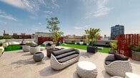 Located in Philadelphia's historic Old City neighborhood, this generously sized rooftop terrace offers an exceptional outdoor experience above the city. The space is thoughtfully divided into distinct zones, combining large-format stone tile patio areas with lush artificial turf lawns that create a vibrant, park-like atmosphere high above street level. Throughout the terrace, an extensive collection of planters — ranging from sculptural round vessels to tall rectangular containers — showcases a variety of trees, shrubs, and flowering plants, bringing rich greenery to the urban setting. Multiple seating arrangements are distributed across the space, featuring contemporary outdoor sofas, lounge chairs, accent chairs, and sun loungers, providing versatile areas for relaxation and social gatherings. A built-in outdoor kitchen with grilling stations supports al fresco dining and entertaining. Decorative lattice privacy screens add architectural interest while defining areas within the open layout. The terrace is crowned by sweeping, unobstructed panoramic views of the Philadelphia skyline to the west and surrounding Old City rooftops, including a striking historic clock tower. Open to abundant natural light and sky, this rooftop destination offers a compelling blend of sophisticated outdoor design and an unmistakable sense of place within one of Philadelphia's most storied districts.