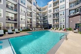 Located at 230 Bill Kennedy Way SE in Atlanta's Grant Park/Ormewood Park area, this outdoor amenity space showcases a thoughtfully designed courtyard at the heart of a modern multi-story residential community. The centerpiece is a generously sized resort-style swimming pool with crystal-clear blue water, offering both shallow entry points and traditional pool access via stainless steel railings. The pool deck is lined with contemporary lounge chairs, providing ample space for sunbathing and relaxation. Surrounding the pool, carefully curated landscaping features ornamental grasses, shrubs, boulders, and seasonal plantings that add natural texture and color to the hardscaped environment. The courtyard is enclosed on multiple sides by the building's striking exterior, which combines gray lap siding, dark navy accents, and warm brick facades with large windows and private resident balconies overlooking the amenity below. A shaded pergola structure and additional seating areas offer comfortable outdoor gathering spaces. The entry area features cozy wicker lounge seating nestled within a brick alcove, creating an inviting welcome. The overall ambiance is vibrant yet relaxed, blending urban architecture with resort-inspired outdoor living in the heart of Atlanta.