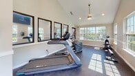 Located at 2950 E North St in Greenville, South Carolina, this fitness center offers a well-equipped, single-floor workout facility designed to support a comprehensive exercise routine. The space features vaulted ceilings with recessed lighting and a ceiling fan, creating an open and airy atmosphere throughout.

Large multi-pane windows line the walls, flooding the interior with generous natural light and offering views of the surrounding outdoor area, including a pool and lush tree canopy. A glass-paneled door provides direct access to an exterior deck or patio, further connecting the fitness space to the outdoors.

The facility is outfitted with a full suite of cardio equipment, including multiple treadmills and an elliptical trainer, as well as a dedicated strength training area featuring a Precor cable machine, a weight bench, and a tiered dumbbell rack with a range of free weights. Wall-mounted mirrors along one side of the room enhance the sense of space and allow for proper form monitoring during workouts. A wall-mounted television is also present for entertainment during exercise sessions.

The durable rubber flooring throughout is well-suited for high-traffic fitness use, providing comfort and safety underfoot. The neutral-toned walls and clean architectural lines give the space a modern, functional aesthetic, making it an inviting environment for daily fitness activity.
