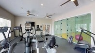 Located at 300 Woodway Bluff Cir in Cary, NC, this single-floor fitness center offers a well-equipped workout facility designed to support a wide range of exercise needs. The space is divided into distinct zones, seamlessly integrating cardio training, strength equipment, and court sports into one cohesive environment.

The cardio area features multiple treadmills, elliptical machines, and a stationary recumbent bike, accompanied by a wall-mounted flat-screen television for entertainment during workouts. Adjacent to the cardio zone, a dedicated strength training area houses a comprehensive array of cable machines, weight benches, and selectorized equipment suitable for both upper and lower body training. Stability balls and additional accessories round out the functional fitness offerings.

Natural light floods the space through a generous row of windows lining the walls, creating a bright and energizing atmosphere. Multiple ceiling fans are distributed throughout, ensuring consistent airflow and comfort across the entire floor. The dark rubberized flooring provides both durability and safety underfoot.

A standout feature of the facility is the glass-enclosed racquetball court, visible from the main workout floor through full-height glass panels, adding a dynamic visual element and an additional athletic option. Full-length mirrors along one wall further enhance the sense of openness. The textured ceiling and neutral wall tones complete the clean, functional aesthetic of this comprehensive fitness facility.