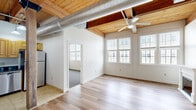 Located at 51 Maple St in Rockland, MA, this 2-bedroom, 2-bathroom unit is defined by its distinctive architectural character. A standout feature throughout is the warm, exposed wood beam ceiling with tongue-and-groove planking, complemented by ceiling fans that enhance the airy, loft-inspired atmosphere. The living room showcases light hardwood flooring, a classic white-mantled fireplace, and a wall of windows dressed with blinds that flood the space with natural light. The kitchen is well-appointed with warm wood cabinetry, dark granite countertops, stainless steel appliances including a range, microwave, dishwasher, and refrigerator, and tile flooring — anchored by a striking exposed structural wood post that nods to the building's heritage. Both bedrooms continue the exposed beam ceiling theme, featuring plush carpeting, generous windows with outdoor views, and ceiling fans. The primary bedroom offers a spacious layout with ample room for furnishings. The two bathrooms are clean and functional, each featuring a full tub-shower combination and tile flooring. Two closets provide practical storage throughout. The unit offers a cohesive blend of historic character and modern updates.