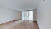 This single-floor residence offers a clean, functional layout designed for comfortable everyday living. The main living area is a standout feature, characterized by its generous open floor plan, neutral-toned wall-to-wall carpeting, and crisp white walls that create a bright, airy atmosphere. Large sliding glass doors span a significant portion of one wall, flooding the interior with natural light while providing direct access to an outdoor area. Vertical blinds offer adjustable light control and privacy. Floor-level air vents are integrated discreetly into the space, maintaining the room's clean aesthetic. The smooth white ceiling completes the open and uncluttered feel of the interior. The overall design is simple and versatile, providing a neutral canvas well-suited to a variety of furnishing styles. The abundance of natural light and the connection to the outdoors through the sliding glass doors contribute to a welcoming and spacious ambiance throughout the living space.