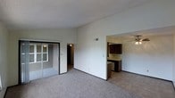 This single-floor space offers an open and functional layout designed for comfortable everyday living. The main living area is finished with wall-to-wall carpet in a neutral tone, creating a warm and cohesive feel underfoot. A sliding glass door provides access to an exterior area, allowing natural light to filter into the space while offering a convenient outdoor connection.

The living area flows seamlessly into the kitchen, which is partially open and visible through a wide pass-through opening. The kitchen features dark wood cabinetry that contrasts with lighter countertops and is equipped with a ceiling fan with integrated lighting, adding both airflow and illumination to the space. The kitchen flooring transitions to a hard surface, distinguishing it from the carpeted living area.

A hallway leading from the main living space provides access to additional rooms beyond. The walls throughout are finished in a clean, neutral white, complementing the textured ceiling treatment. An overhead HVAC vent is positioned centrally, ensuring consistent climate control. Overall, the space presents a straightforward, practical layout with clear zone separation between living and kitchen areas.