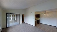 This single-floor space offers an open and functional layout designed for comfortable everyday living. The main living area is finished with wall-to-wall carpet in a neutral tone, creating a warm and cohesive feel underfoot. A sliding glass door provides access to an exterior area, allowing natural light to filter into the space while offering a convenient outdoor connection.

The living area flows seamlessly into the kitchen, which is partially open and visible through a wide pass-through opening. The kitchen features dark wood cabinetry that contrasts with lighter countertops and is equipped with a ceiling fan with integrated lighting, adding both airflow and illumination to the space. The kitchen flooring transitions to a hard surface, distinguishing it from the carpeted living area.

A hallway leading from the main living space provides access to additional rooms beyond. The walls throughout are finished in a clean, neutral white, complementing the textured ceiling treatment. An overhead HVAC vent is positioned centrally, ensuring consistent climate control. Overall, the space presents a straightforward, practical layout with clear zone separation between living and kitchen areas.